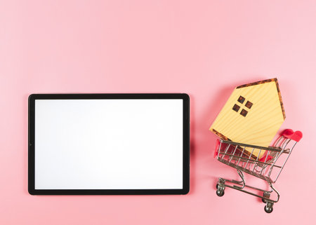 Top view or flat layout of digital tablet and wooden house model in shopping trolley  on pink  background with copy space, home purchase concept.の写真素材