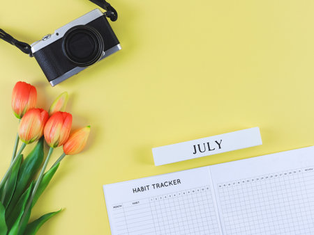 Top view or flat lay of habit tracker book, wooden calendar July, digital camera and red yellow tulips flowers on yellow background with copy space.の写真素材