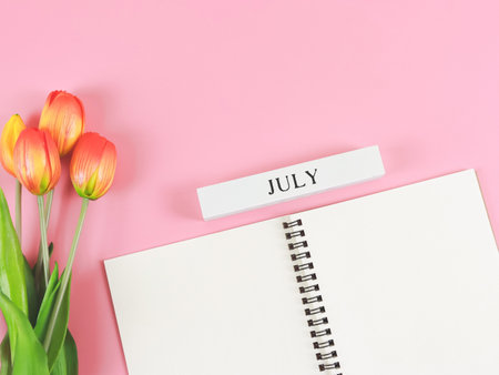 Top view or flat lay of opened diary or notebook with wooden calendar july  on pink background with orange-yellow tulips.の写真素材