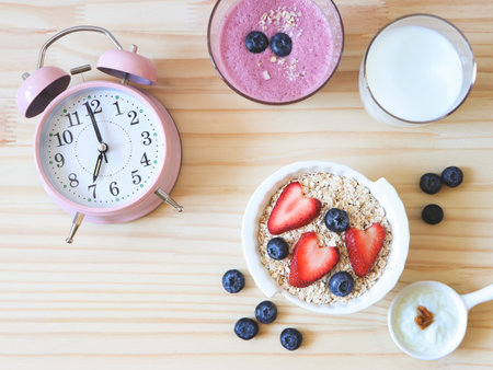 Top view or flat lay of alarm clock, breakfast with oat or granola in white bowl, fresh blueberries, strawberries, a  glass of milk, blueberry smoothie  and yogurt on wooden table. Healthy breakfast concept.の写真素材