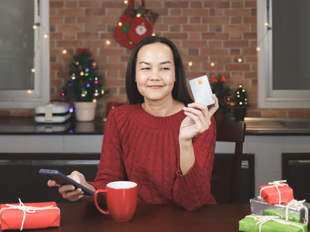 Portrait of Asian woman wearing red  sweater sitting  with red cup of coffee  and gift boxes in the kitchen with Christmas decoration, holding mobile phone, smiling and looking at credit card in her hand.の写真素材