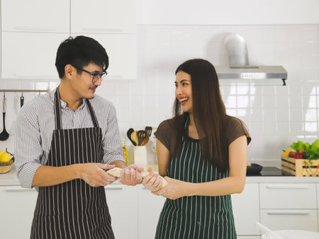 Happy Asian man wearing eye glasses preparing bakery in modern kitchen with beautiful woman with...