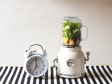 Front view of  white vintage blender or smoothie maker machine  with vegetables and fruit inside and white vintage alarm clock shows 7 o'clock  on  black and white stripe table cloth and white wall.の写真素材