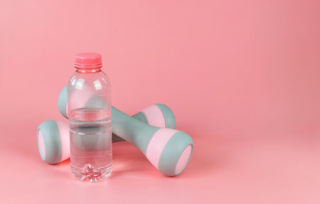 Front view of pink bottle of water and two dumbbells isolated on pink background. Health and fitness concept.の写真素材
