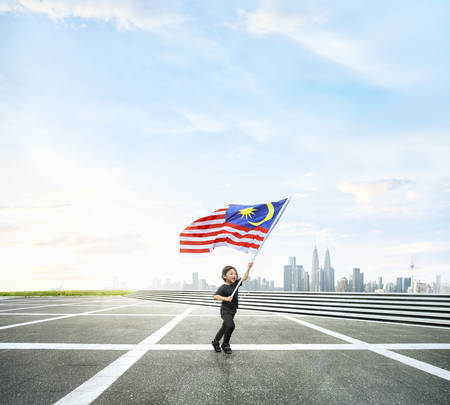 Happy boy running while holding Malaysia flag on open space with Kuala Lumpur cityscape.の写真素材