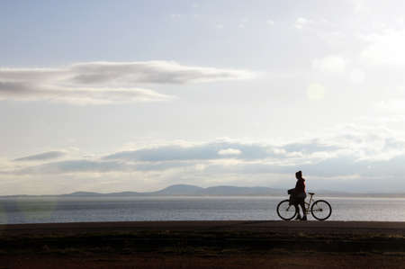 A woman walking with her bicycle on the side of the path. In the background you can see the sea and some mountainsの写真素材