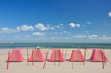 Red and white beach tents on an almost deserted beach, with a calm sea. Some people sunbathe on the sandの写真素材