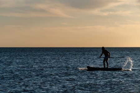 A man is paddling a surfboard in the ocean. The sky is a mix of orange and blue, creating a serene and peaceful atmosphereの写真素材