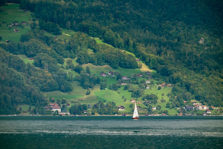A sailboat is sailing on a lake near a town. The sailboat is small and white. The lake is calm and the water is clear. The town is surrounded by trees and hills. The scene is peaceful and sereneの写真素材