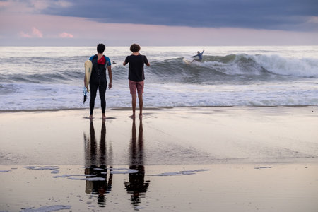 Two men standing on the beach, one holding a surfboard. The other man is watching a surfer ride a wave. Scene is relaxed and peaceful, as the two men enjoy the beach and the oceanの写真素材