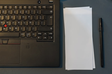 Close-up of a laptop keyboard, blank paper, and pen on a dark blue desk.