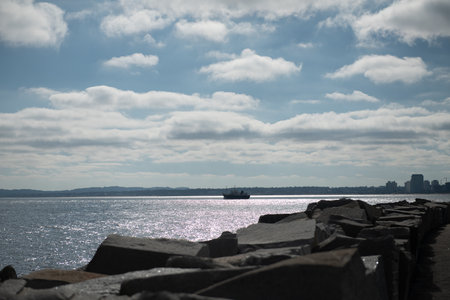 Ship sailing on calm sea under partly cloudy sky with rocky shoreline foregroundの写真素材
