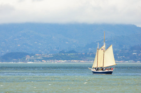 Classic sailboat in scenic bay with lush hills and coastal cloudsの写真素材