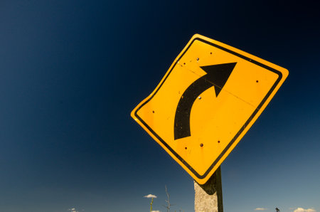 Yellow road sign against blue sky warns of slight curve aheadの写真素材