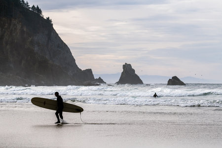 A person is walking on the beach with a surfboard. The sky is cloudy and the water is choppyの写真素材