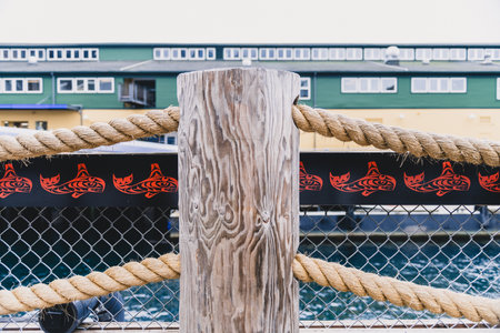 A wooden post with a rope tied to it and a black and red design on it. The fence is behind the post and the post is near a body of waterの写真素材