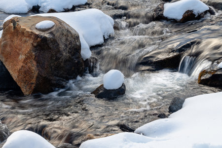 A stream of water flows past a large rock covered in snow. The scene is peaceful and serene, with the snow covering the ground and the water flowing calmlyの写真素材