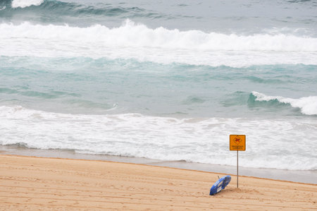 Beach with warning sign, lifeguard gear, and ocean waves crashing ashoreの写真素材
