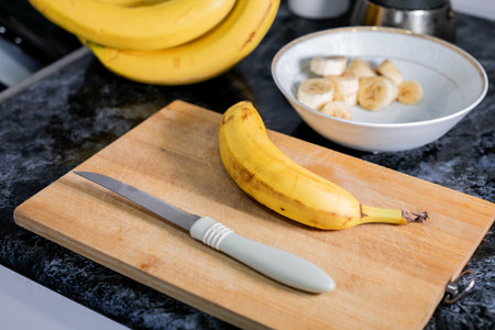 A ripe banana alongside a kitchen knife on a wooden cutting board, with sliced bananas in a bowl in the background, suggesting healthy eating or food preparationの写真素材