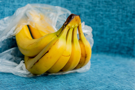 A bunch of ripe bananas in a plastic bag on a textured blue background, depicting the concept of food packaging and preservationの写真素材