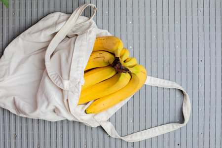 A bunch of ripe bananas in a white cotton tote bag on a grey striped background, suggesting sustainable grocery shopping or zero waste lifestyle conceptの写真素材