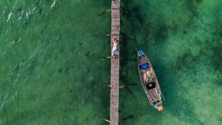 Aerial view of a person lying on a wooden pier next to a small boat in clear turquoise waters, conveying a sense of relaxation and solitudeの写真素材