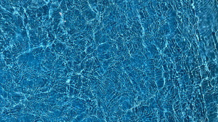 A close up texture of sunlit, clear blue swimming pool water rippling gently, ideal for summer or...