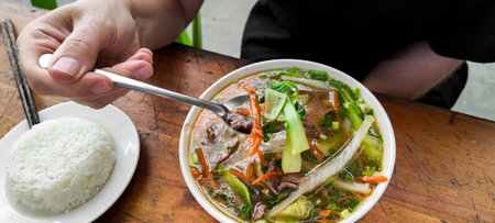 Person eating traditional Asian beef and vegetable soup with a side of white rice, concept of everyday dining experienceの写真素材