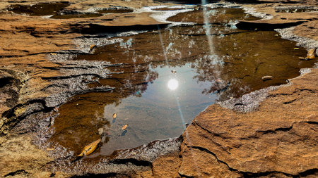 A serene puddle reflecting the bright sun amidst rugged brown rocks, conveying a concept of natural tranquility and the simplicity of earths beautyの写真素材