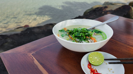 A fresh bowl of Vietnamese pho served with lime and chili on a wooden table with a serene water background, representing traditional cuisine or healthy eatingの写真素材