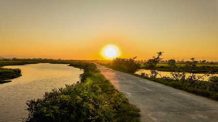 Breathtaking sunset over a serene river with lush greenery, featuring a tranquil rural pathway, perfect for backgrounds with space for textの写真素材