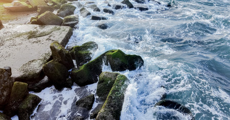 Mossy ocean boulders with dynamic waves crashing against them at sunset, ideal for backgrounds and...