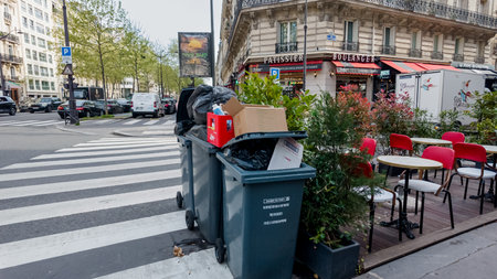 Overfilled city trash bins on a Parisian street with outdoor cafe seating nearby, depicting urban...