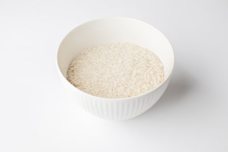 A ceramic bowl filled with uncooked white rice on a white background, optimal for illustrating concepts of basic nutrition or Asian cuisine preparationの写真素材