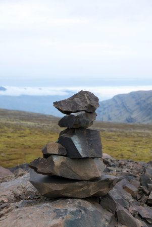 A cairn of dark stones stands against a backdrop of landscape and cloudy sky.