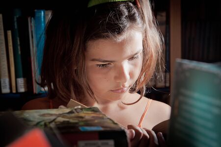 A little girl reading in a library, completely absorbed in her book, seen through the bookshelves.の写真素材
