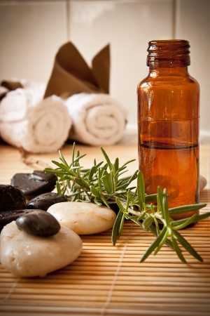 A bottle of aromatherapy oil with herbs, massage stones and towels in the background の写真素材