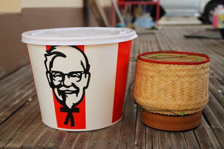 BANGKOK, THAILAND- FEBRUARY 28, 2020 : Crispy fried chicken bucket. KFC is popular fast food chain known as Kentucky Fried Chicken and Glutinous rice boxes in northeastern Thailand Kratib rice put on the wooden table , show olden it, Rice box put sticky rのeditorial素材