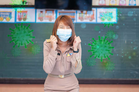 Female thai teacher in uniform standing in front of the classroom wearing mask and greeting with green virus around herの写真素材