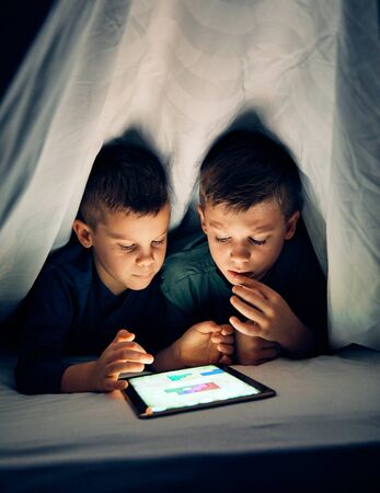School age brothers having fun with a tablet together hiding under sheet at homeの写真素材