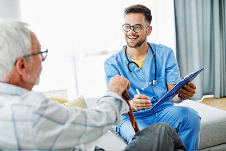 Doctor or male caregiver with senior man holding a cane on sofa at home or nursing home