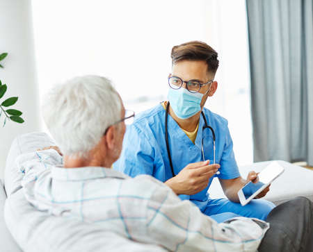 Doctor or nurse caregiver wearing a protective mask helping senior man with a tablet at home or...