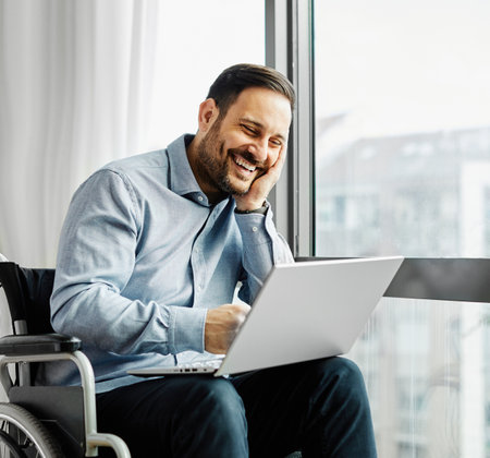 Portrait of a young man in wheelchair using laptop by the window at home or in hospitalの写真素材