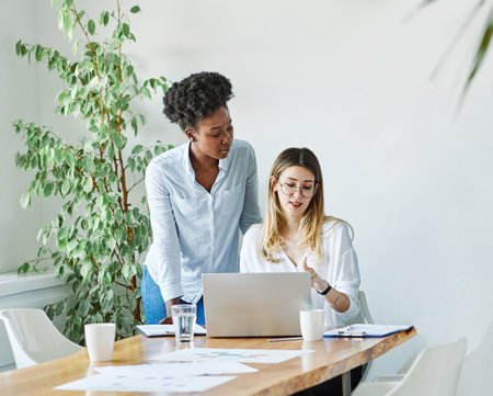 Portrait of two of young businesswomen multiethnic working with laptop on desk and talking in a start up officeの写真素材