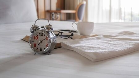 Alarm clock, book, cup of coffee and accessories on white bedding sheets with copy space. Morning...