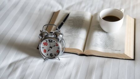 Alarm clock, book, cup of coffee and accessories on white bedding sheets with copy space. Morning lifestyle concept.の写真素材
