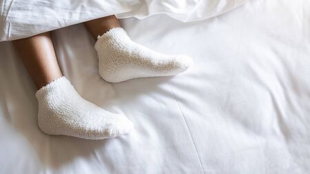 Woman feet in warm woolen socks on white bedclothes. Close up of young woman lying in bed at home....