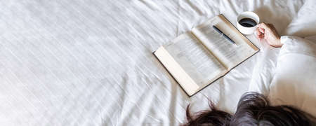 Time to drink coffee. Asian lazy woman lying and holding a cup of coffee on the bed after read a book in the morning with soft morning light. Morning lifestyle concept.の写真素材