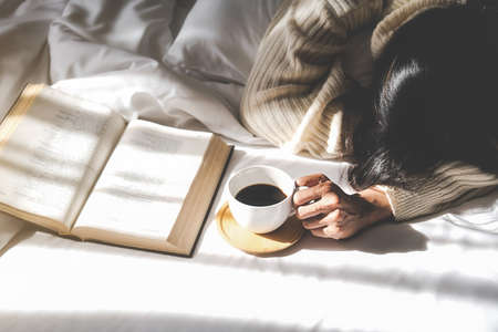 Time to drink coffee. Asian lazy woman lying and holding a cup of coffee on the bed after read a book in the morning with soft morning light. Morning lifestyle concept.の写真素材