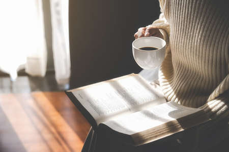 Asian woman holding cup of coffee and reading a book beside the window after get up in morning....